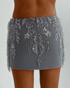 A close-up of a model wearing 'The Alice- tassel skirt' mini silver skirt heavily embellished with silver sequins and beadwork, showing the back design and side fringe details. 