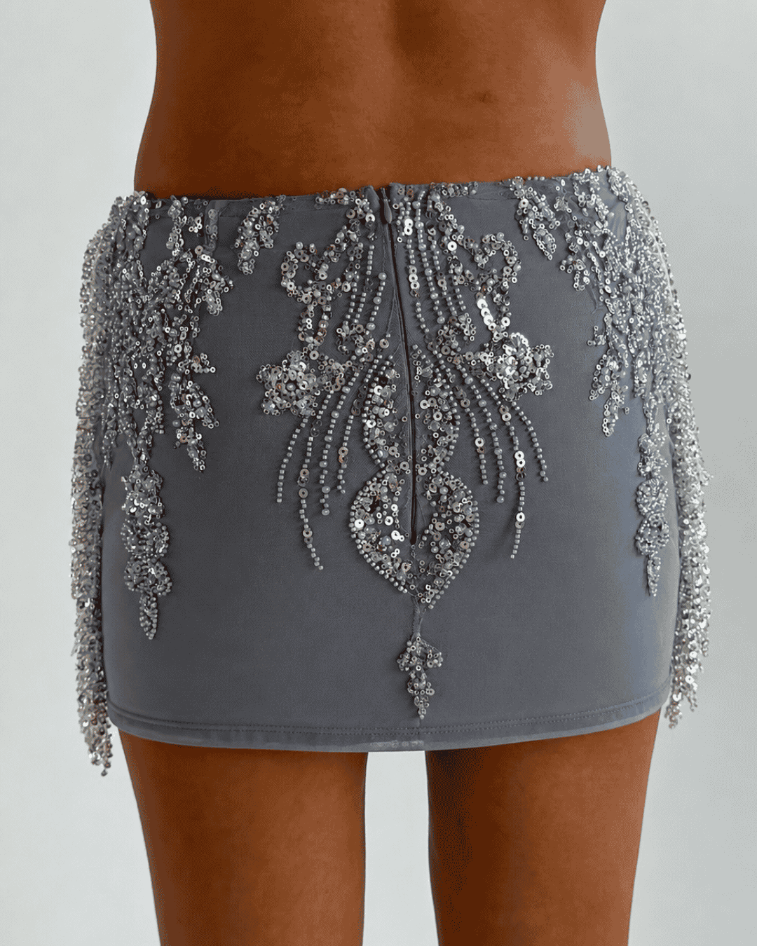 A close-up of a model wearing 'The Alice- tassel skirt' mini silver skirt heavily embellished with silver sequins and beadwork, showing the back design and side fringe details. 